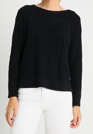 Femme portant un pull en maille texturée noire à manches longues, assorti d'un pantalon blanc, vue des épaules aux hanches.