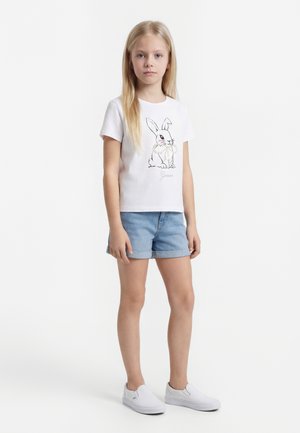Meisje met lang blond haar dat een wit T-shirt met een konijnengrafiek draagt, lichtblauwe denimshorts en witte slip-on schoenen, staand en recht vooruit kijkend.
