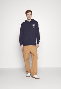 Hanorac cu glugă bleu marin, cu buzunar frontal și design grafic; asortat cu pantaloni bej lejeri și adidași albi.