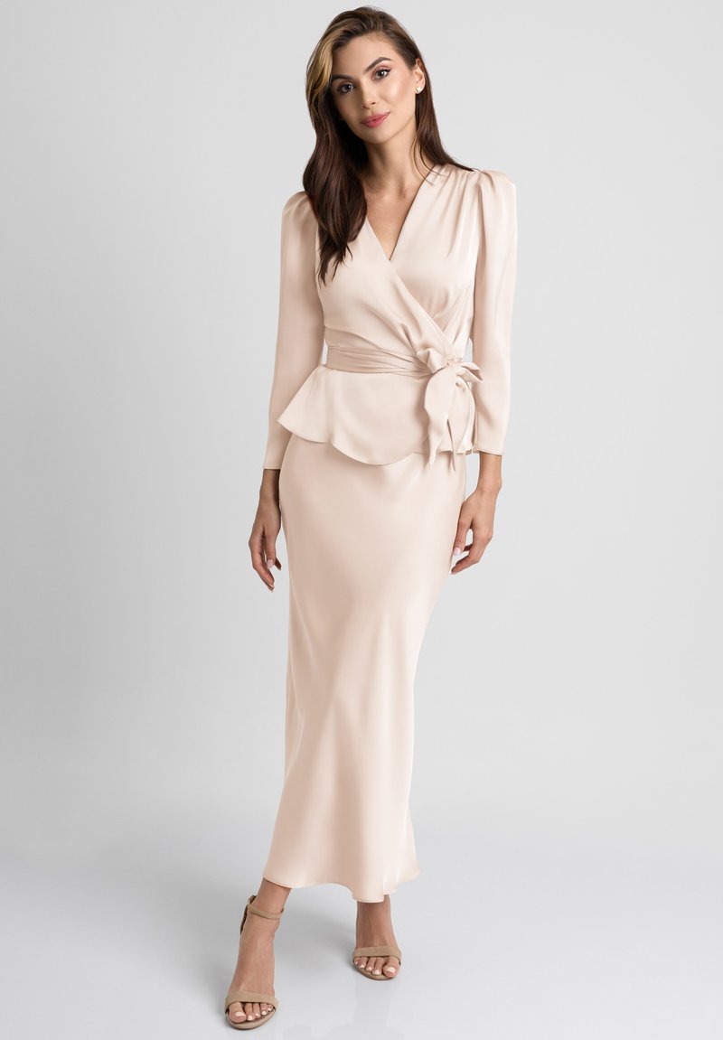 Długa, kremowa sukienka w stylu narzutki z długimi rękawami, peplum w talii, gładką teksturą i dekoracyjnym wiązaniem w talii. Całość dopełniają wysokie obcasy.