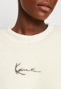 Mikina z krémově zbarveného fleecu s texturovaným povrchem a žebrovaným límcem. Na přední straně je kovový akcent s logem.