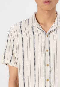 Camicia a maniche corte con bottoni di colore crema, strisce verticali navy, tessuto testurizzato e bottoni marroni. Design del colletto moderno.