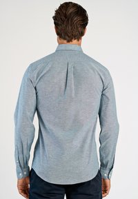 Ανοιχτό μπλε πουκάμισο με μακριά μανίκια, γιακά με κουμπιά, πτυχώσεις στο πίσω μέρος και μανσέτες με κουμπιά. Κατασκευασμένο από υφασμάτινη ύφανση με διακριτικό μοτίβο.