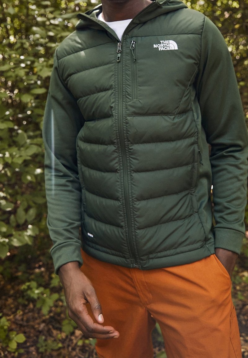 Vihreä pehmustettu takki, jossa on vetoketjukiinnitys, huppu ja logo rinnassa; sisältää sileää kangasta ja teksturoitua paneelia; käytetään oranssien housujen kanssa.