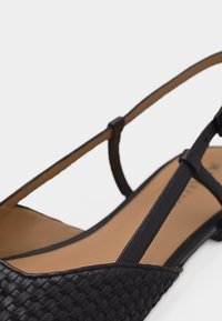Μαύρο πλεκτό δερμάτινο επίπεδο σανδάλι με μυτερή μύτη, λεπτό λουράκι στον αστράγαλο και μπεζ εσωτερική σόλα σε λευκό φόντο.