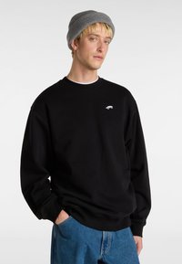 Felpa nera con collo tondo e polsini a costine, caratterizzata da una piccola grafica bianca sul petto. Abbinata a jeans in denim blu.