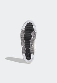 Semelle de chaussure en caoutchouc noir texturé, avec des accents gris et un bord blanc. Présente un motif hexagonal et des points de traction circulaires.