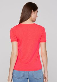Camiseta de manga corta en color coral con cuello redondo y una tela suave y texturizada, llevada con jeans azules, mostrando una vista trasera.