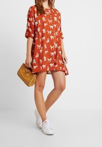 Rotes Kleid mit floralem Muster und schwarzen Punkten, mit kurzen Ärmeln und lockerer Passform. Eine geflochtene Handtasche und weiße Sneaker runden den Look ab.