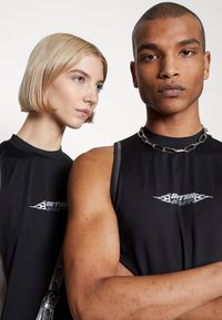 Zwei Models tragen enganliegende schwarze ärmellose Oberteile mit einem grafischen Logo und einer Kettenhalskette. Der Hintergrund ist schlicht weiß und lenkt den Fokus auf ihre Outfits.