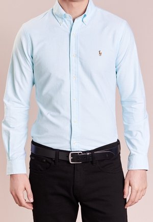 Homme portant une chemise bleu clair à boutons et un pantalon noir avec une ceinture noire, debout contre un fond neutre.