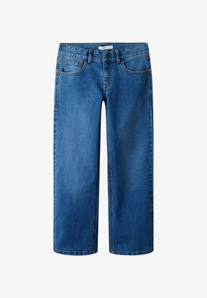 Niña joven con cabello largo y castaño, vistiendo una camiseta blanca con la palabra "Botanical", jeans azules y zapatos marrones, sobre un fondo liso de color gris claro.