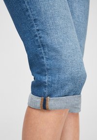 Jean en denim bleu clair avec des poignets retroussés, présentant des coutures contrastantes et une texture légèrement décolorée. Gros plan sur la zone de la jambe.