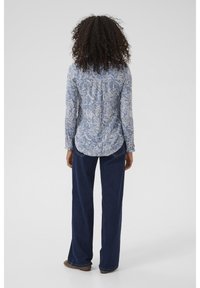 Chemise bleue à motif avec col boutonné, ornée de dessins floraux, portée avec un pantalon large bleu foncé et des chaussures marron.