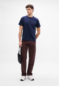 Maglietta in cotone blu navy con collo a giro, abbinata a pantaloni marrone scuro e sneaker bianche, mentre tiene una borsa a tracolla nera.