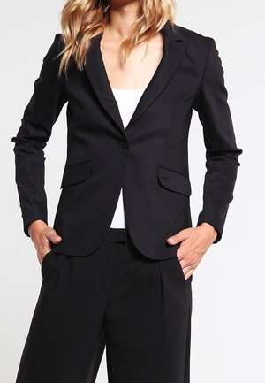 Kobieta ubrana w dopasowaną czarną marynarkę na białym topie oraz pasujące czarne szerokie spodnie, ręce w kieszeniach, stojąca na jednolitym tle.