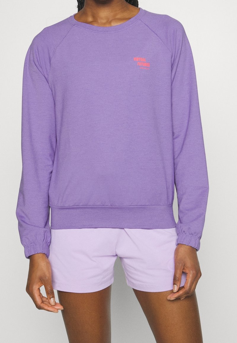 Personne portant un sweat-shirt violet à manches longues avec un petit texte rouge et un short violet clair, debout devant un fond uni.