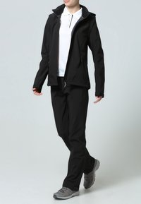 Chaqueta de softshell negra con un diseño ajustado, cremallera frontal y capucha, combinada con pantalones negros a juego y zapatos de senderismo grises.