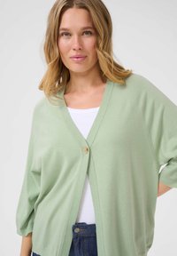 Cardigan vert clair en matériau doux, avec un col en V, une coupe décontractée et une fermeture à un bouton, porté sur un haut blanc.