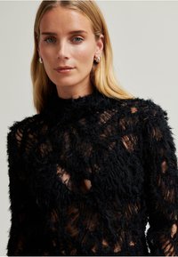 Černý krajkový top s třepící texturou, vysokým límcem a průhlednými výřezy. Dlouhé rukávy s propracovanými designovými detaily.