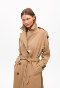 Beige trenchcoat med bälte i midjan, dubbelknäppta knappar och hög krage. Tillverkad av ett slätt tyg, med axelremmar.