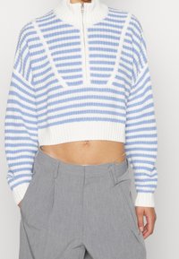 Maglione corto a righe blu e bianche con cerniera a un quarto, abbinato a pantaloni grigi a vita alta e una catena argento visibile in vita.