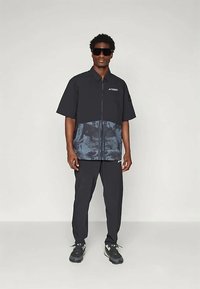 Μαύρο κοντομάνικο μπουφάν με μπλε tie-dye μάκρος και φερμουάρ, σε συνδυασμό με μαύρο παντελόνι και σκούρα αθλητικά παπούτσια. Διακοσμημένο με γυαλία ηλίου.