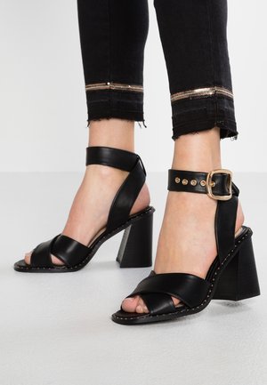Zwarte leren sandalen met hak, voorzien van brede banden, een enkelriempje met gesp en een gestructureerde blokhak. Heeft decoratieve stiksels langs de zool.