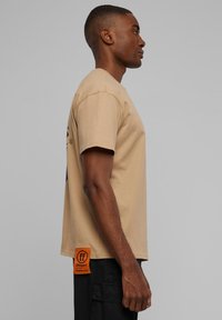 Beige bomulds t-shirt med korte ærmer, der har et grafisk design på forsiden og et orange mærke på sidesømmen.