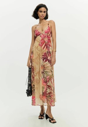 Õlgadeni tumedate juustega naine kannab varrukateta lillelist maksikleiti, hoides musta käsipidurit ja kandes musti kontsaga sandaale.