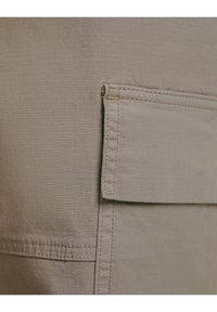 Lähikuva beigeistä kangaslastitaskusta, jossa on läppä ja näkyvät ompelusaumat.