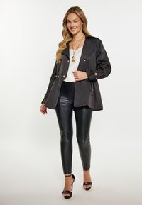 Zwarte lichtgewicht trenchcoat met gouden knopen, gedragen over een witte top en zwarte leren leggings. Open voorkant met strikdetail, hakken.