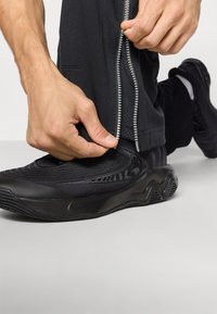 Melni sporta apavi ar teksturētu augšdaļu, izteiksmīgām dizaina zīmēm un gumijas zoli. Modelis pielāgo rāvējslēdzēju uz saskaņotām tumšām biksēm.
