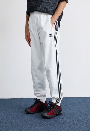 Persoon draagt witte Adidas trainingsbroek met zwarte strepen en zwart-rode sportschoenen, staand op een blauw tapijt.