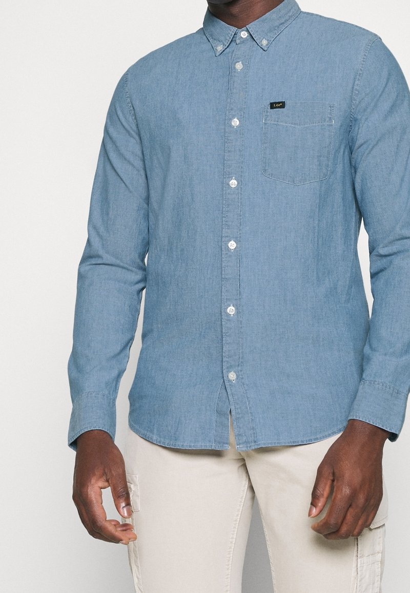 Camicia di jeans azzurro chiaro con colletto abbottonato, taschino anteriore e bottoni bianchi. Caratterizzata da maniche lunghe e orlo dritto.