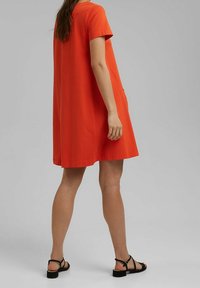 Röd kortärmad klänning med en avslappnad passform, tillverkad av mjukt tyg. Har en rak nederkant och sidofickor. Bärs med svarta sandaletter med remmar.
