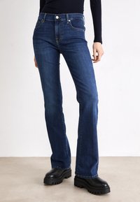 Dark blue denim flared jeans with a mid-rise waist, featuring front pockets, subtle stitching, and a button closure. Worn with black chunky boots.