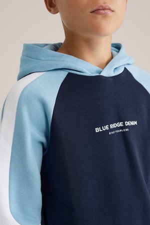 Tengerészeti kapucnis pulóver világoskék ujjakkal és részletekkel. Kangourou zsebével és "BLUE RIDGE DENIM STAY FEARLESS" felirattal ellátva. Puha anyagból készült.