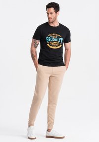 Zwarte katoenen t-shirt met een "BROOKLYN" grafiek in blauw en oranje, gecombineerd met beige joggingbroek en witte sneakers. Eenvoudig en casual ontwerp.