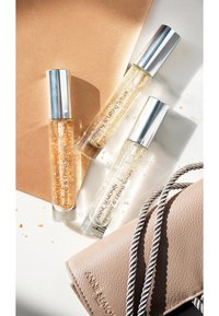 Three clear glass serum bottles with silver caps, containing golden and clear gels, alongside a tan leather wallet with a twisted strap.