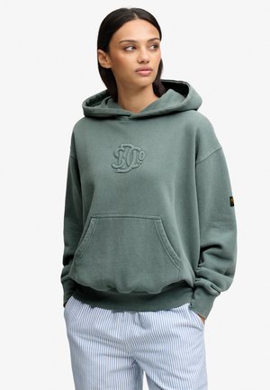 Jeune femme portant un sweat à capuche vert oversize avec poche avant et logo brodé, assorti à un pantalon rayé bleu clair et blanc.