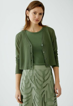 Grüne Strickjacke mit Knöpfen, über einem strukturierten grünen Oberteil und einem passenden Rock mit kunstvollen Ausstanzmustern. Weiche Materialien.