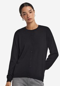 Svart cardigan i mjukt tyg med rund hals, långa ärmar och knappstängning fram. Enkelt mönster och slät textur.