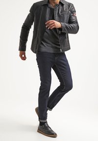 Černá kožená bunda s prošívanou texturou, šedý svetr pod ní, tmavě modré džíny a černé kožené boty s přezkami. Model v pohybu.