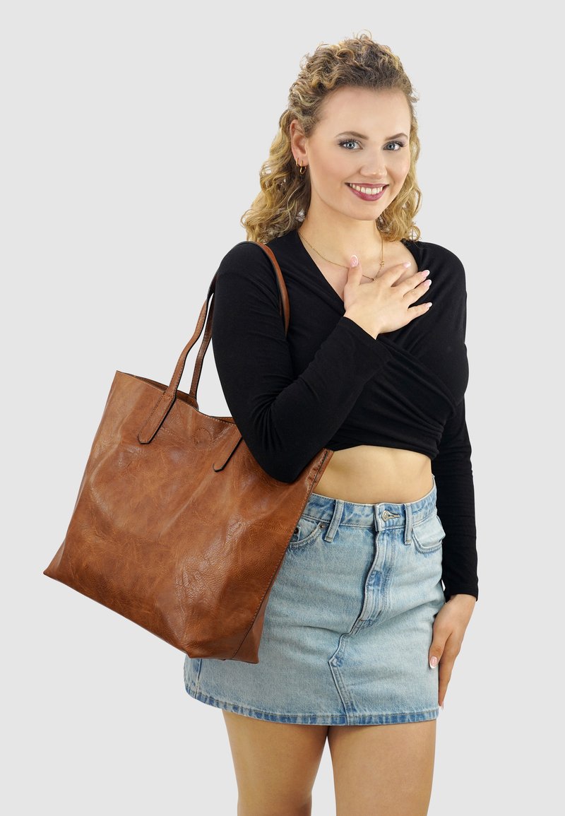 Lachende vrouw met krullend haar draagt een zwart crop top, een denim rok en heeft een grote bruine leren schoudertas.