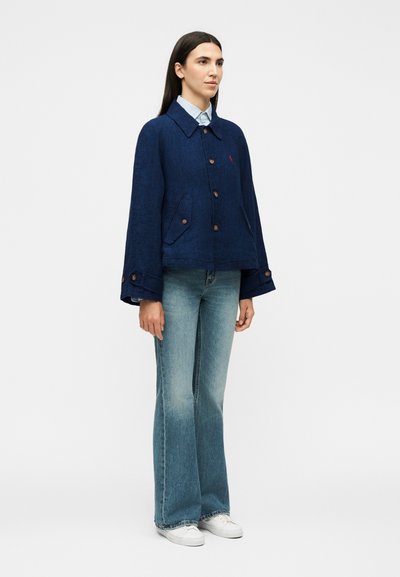 Γυναίκα με μακριά σκούρα μαλλιά που φοράει μια ναυτική μπλε σακάκι με κουμπιά, ανοιχτό μπλε πουκάμισο, καμπάνα τζιν και λευκά αθλητικά παπούτσια, στέκεται μπροστά σε απλό φόντο.