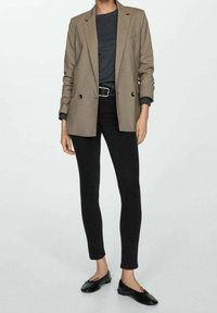 Blazer de doble botonadura en tono beige sobre una blusa gris, combinado con jeans ajustados negros y mocasines negros. Presenta una cintura ceñida con cinturón y mangas arremangadas.