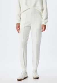 Weiße Sweatpants mit gerippten Bündchen und seitlichen Taschen, die eine glatte Textur und einen leicht lockeren Schnitt aufweisen, kombiniert mit weißen Turnschuhen.