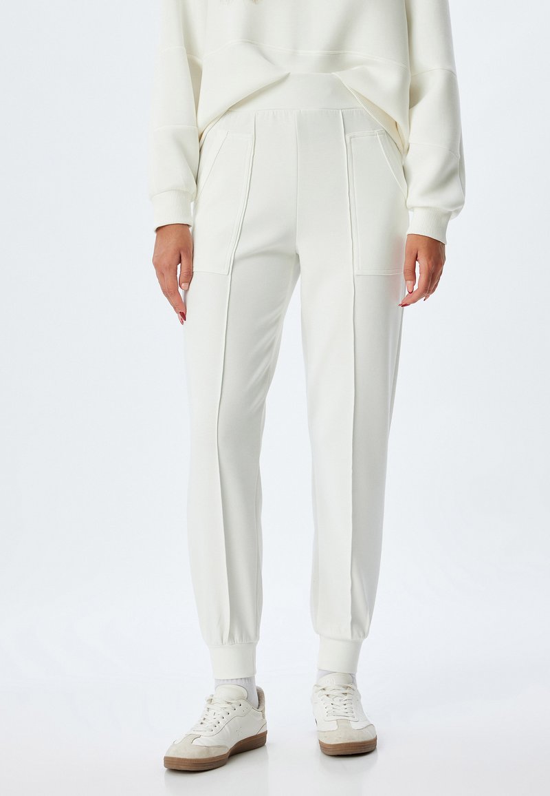 Weiße Sweatpants mit gerippten Bündchen und seitlichen Taschen, die eine glatte Textur und einen leicht lockeren Schnitt aufweisen, kombiniert mit weißen Turnschuhen.