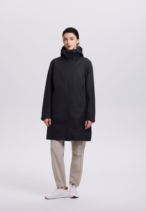 Nainen seisoo päällään pitkä musta hupullinen takki, beigehousut ja valkoiset tennarit yksinkertaisen vaalean taustan edessä.